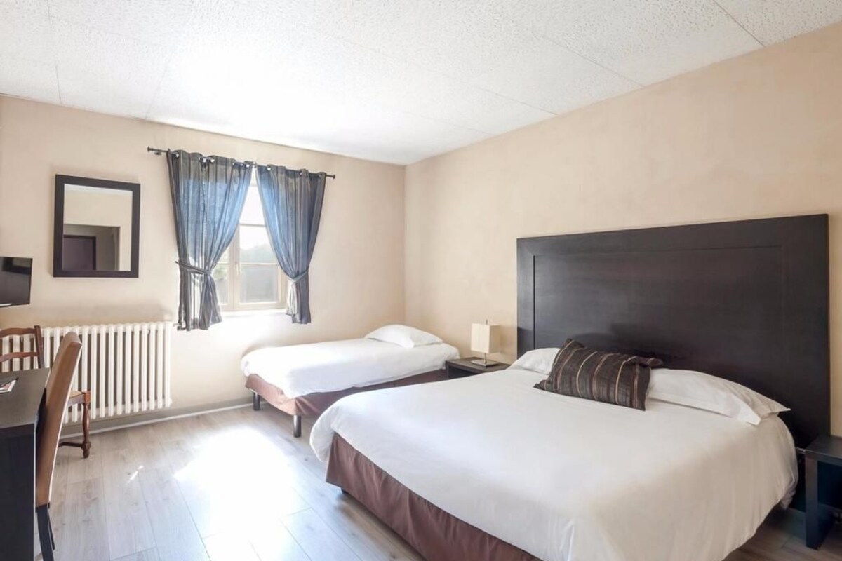 Una habitación de hotel con dos camas individuales, un escritorio, una silla, un radiador y una ventana con cortinas.