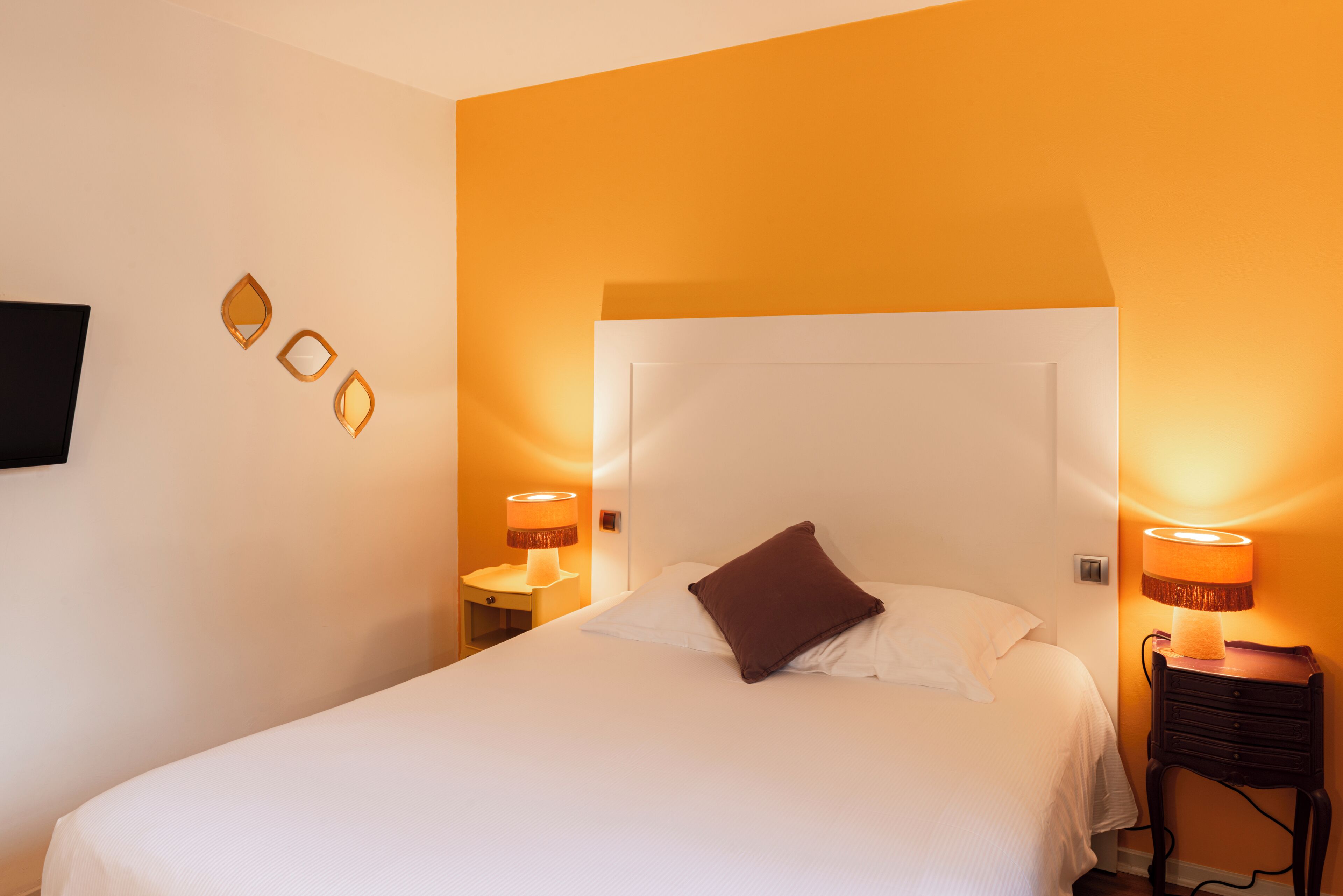 A neatly made bed with a brown pillow, two bedside lamps, and a flat-screen TV mounted on the wall.