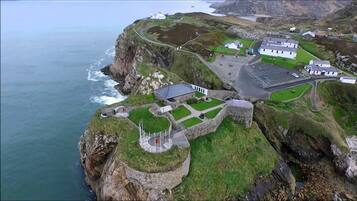 An aerial view of a coastal property with a stone fortification, a parking area, and buildings with a white facade.