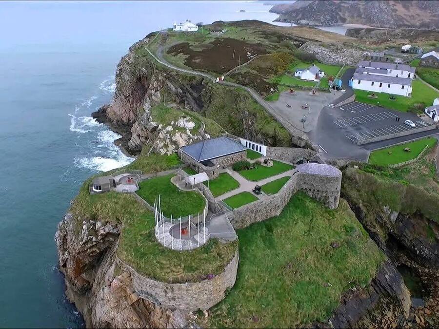 An aerial view of a coastal property with a stone fortification, a parking area, and buildings with a white facade.