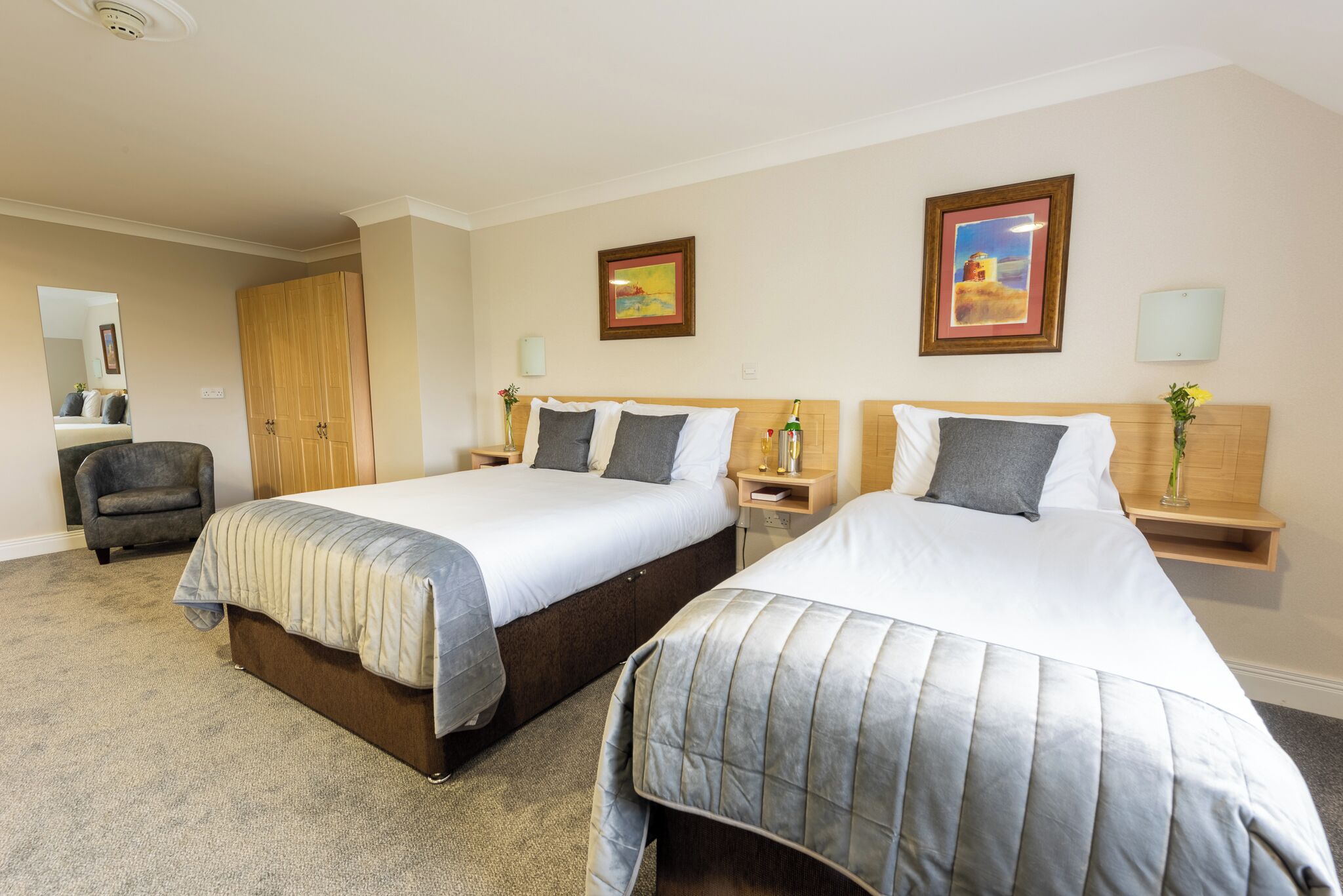 A hotel room with two beds, a chair, a wardrobe, and a mirror.