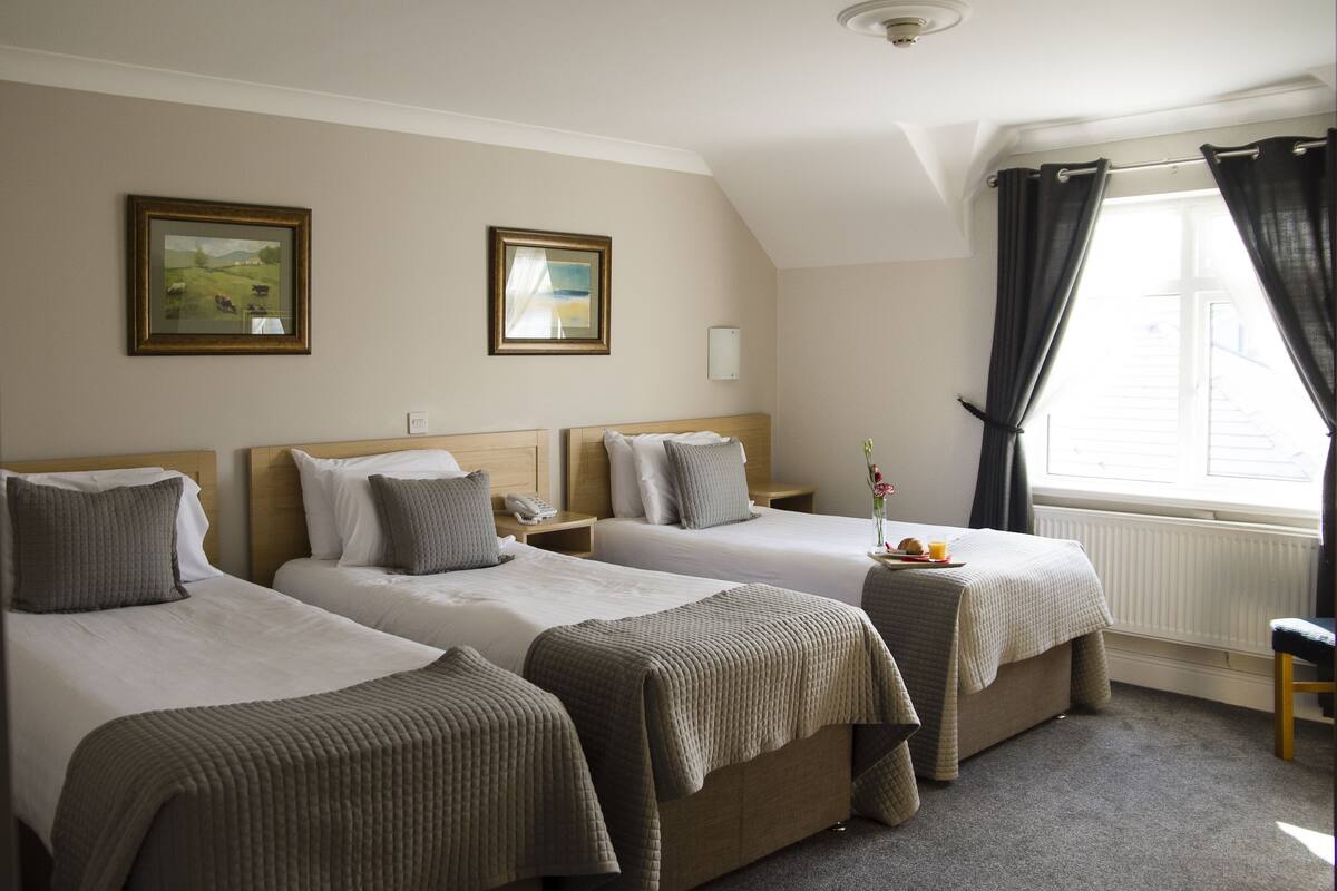 Una habitación de hotel con dos camas individuales, una mesita con un plato de fruta y un ventanal con cortinas.