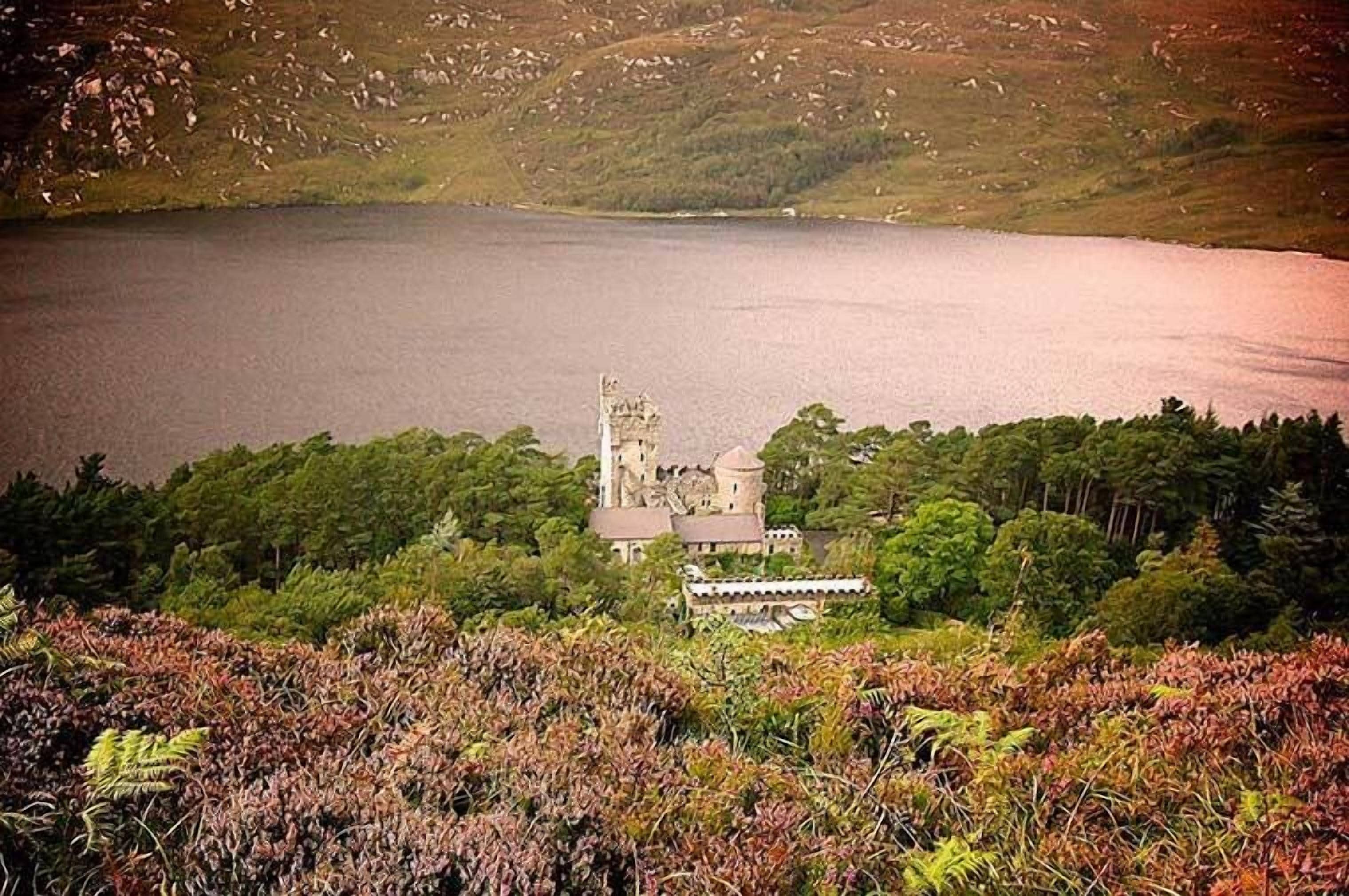 A historic building with a tower, surrounded by dense greenery and a large body of water.