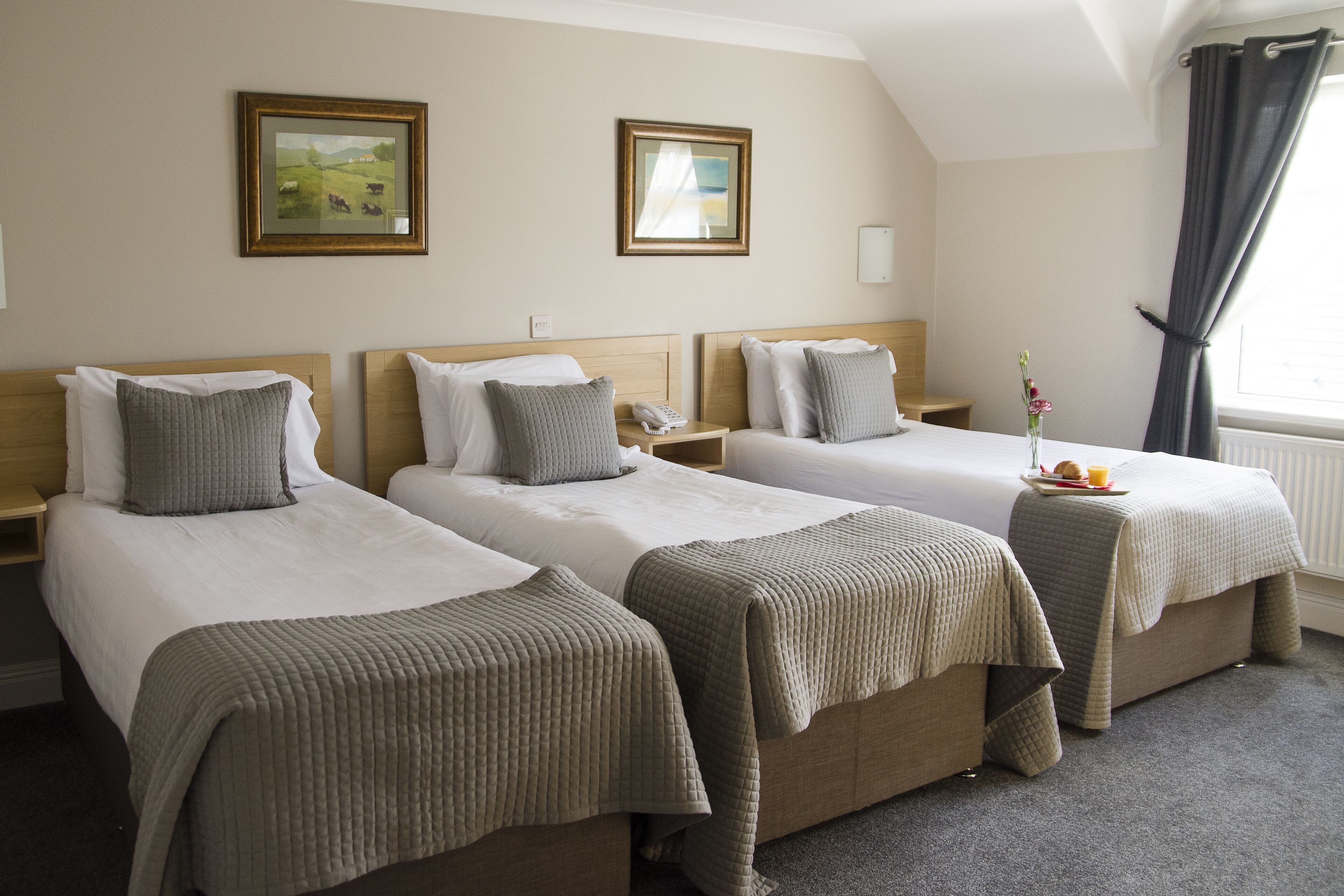 A hotel room with three single beds, each with a pillow and a blanket. There are framed pictures on the wall, a bedside table with a phone, and a window with curtains.