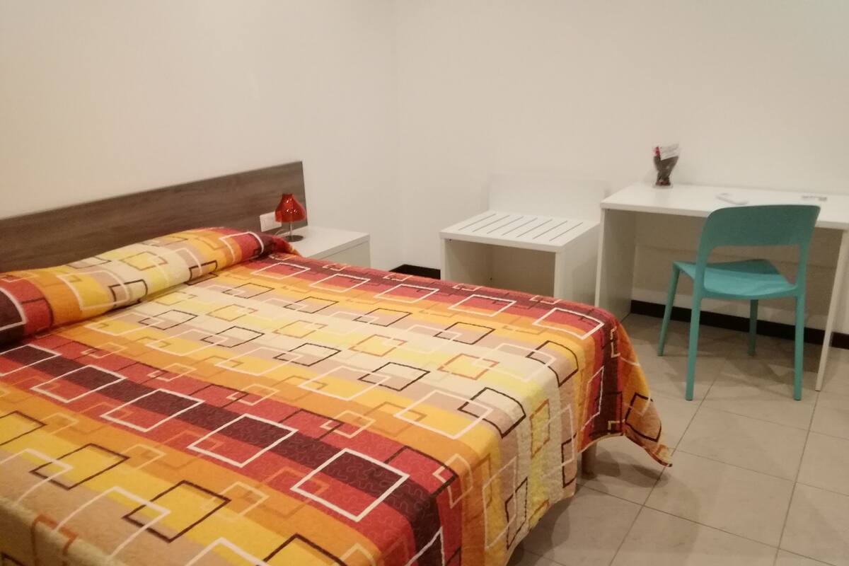 Una cama con un edredón estampado, un escritorio con una silla y una pequeña lámpara roja en la mesita de noche.
