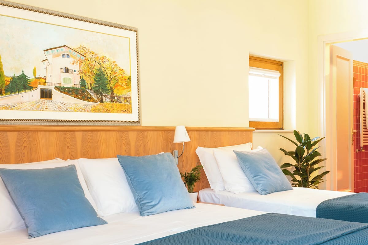 Ein Hotelzimmer mit einem Bett, einem Nachttisch, einer Lampe, einer Topfpflanze und einem Gemälde eines Gebäudes an der Wand.