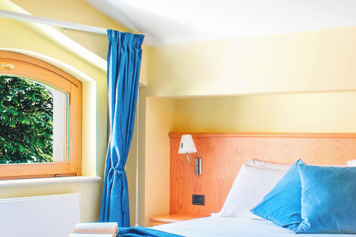 Ein Hotelzimmer mit Bett, Nachttisch, Lampe, einem Fenster mit Vorhängen und Blick ins Grüne.