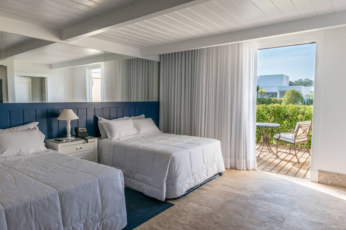 A hotel room with two beds, a nightstand, a lamp, and a view of the outdoor patio.