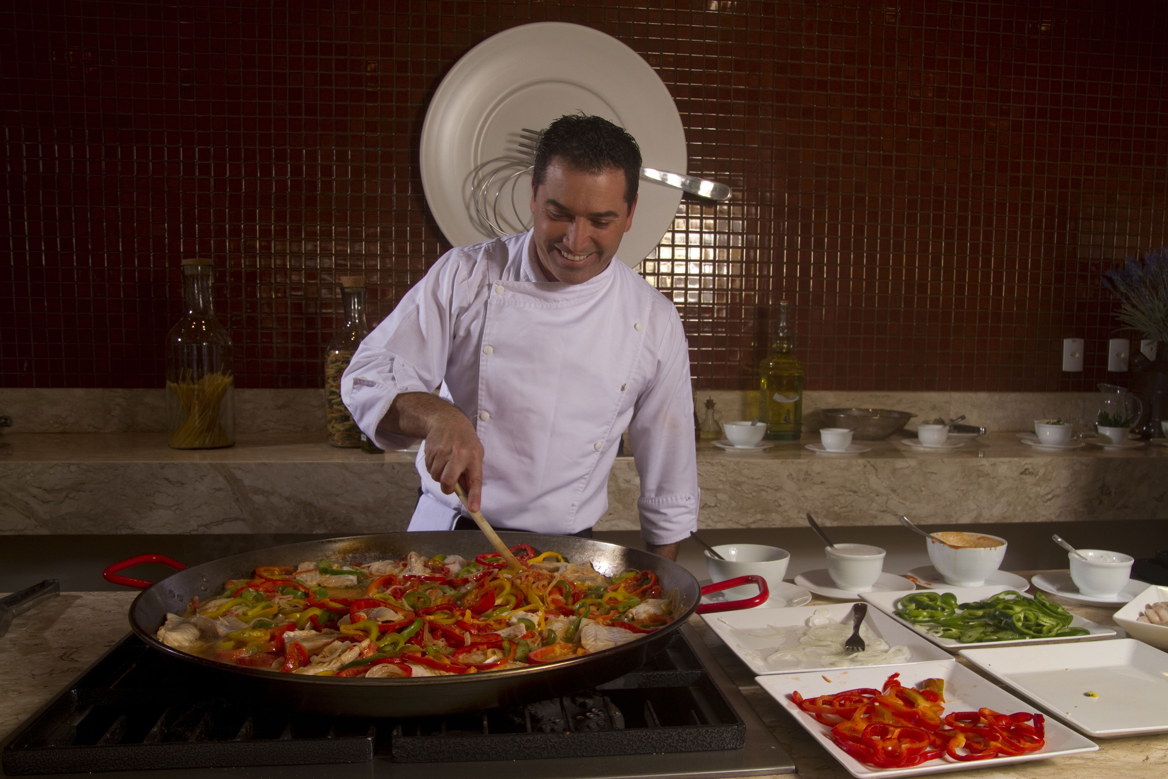 Un cocinero preparando comida en una cocina, con una sartén grande, diversos utensilios de cocina e ingredientes sobre la encimera.
