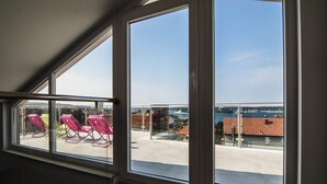 Une pièce dotée de grandes fenêtres donnant sur un patio agrémenté de chaises roses, avec vue sur une étendue d’eau et des bâtiments.