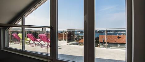 A room with large windows overlooking a patio area with pink chairs and a view of a body of water and buildings.
