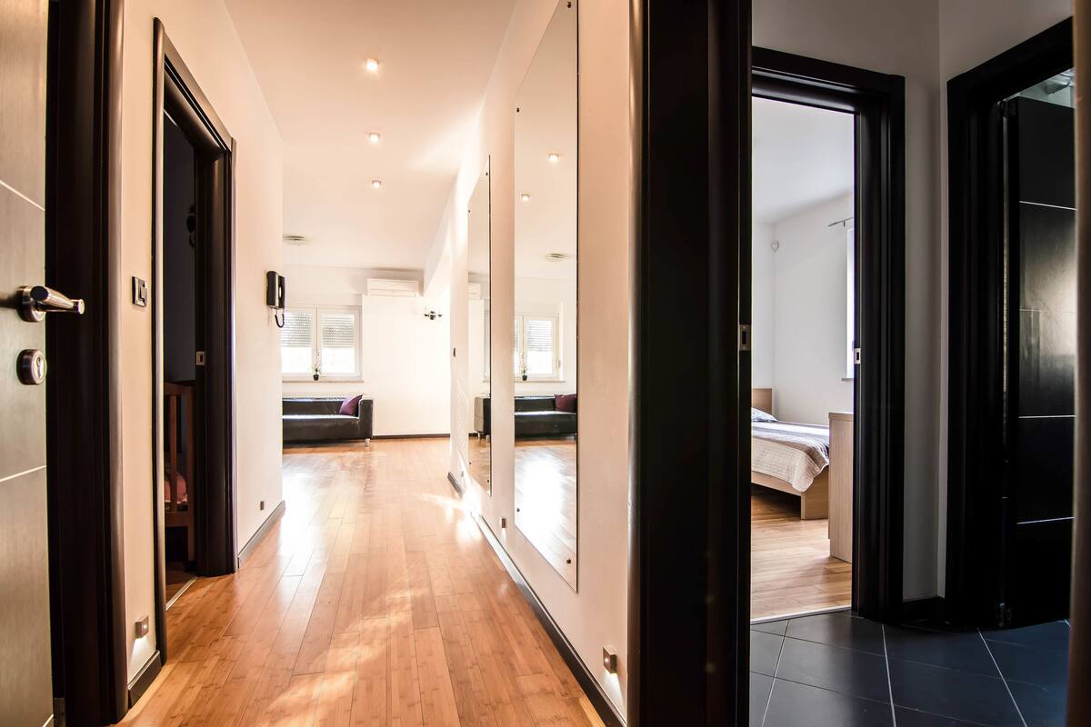 A hallway with wooden flooring, a mirror, and a door leading to a bedroom with a bed and a nightstand.