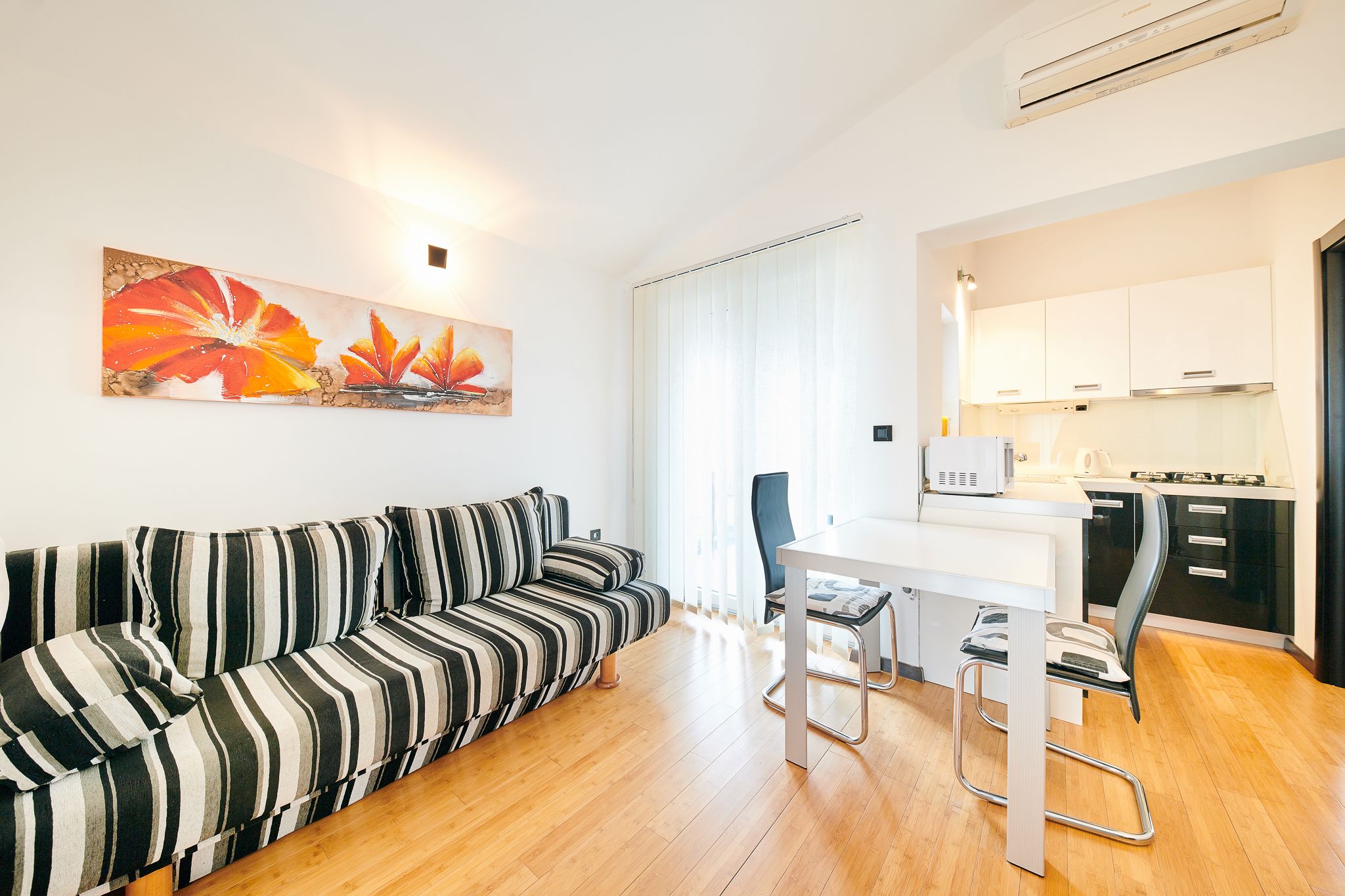 Un salon moderne avec un canapé à rayures, une table à manger avec deux chaises et un coin cuisine doté de meubles blancs et d’une cuisinière.