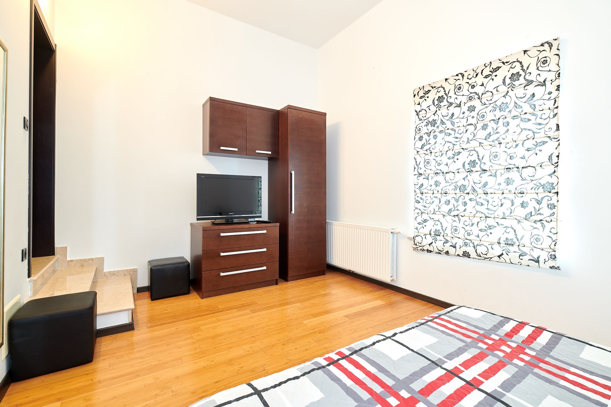 A modern bedroom with a wooden floor, a bed with a patterned blanket, a TV, a wardrobe, and a radiator.