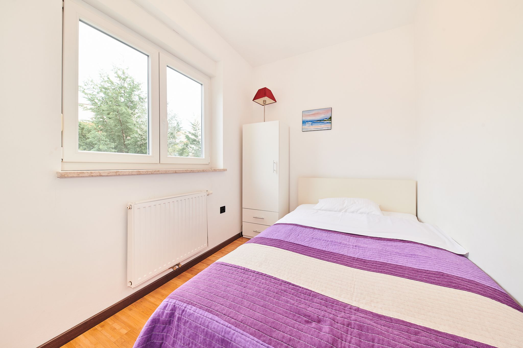 Un dormitorio con cama, ventana, radiador, armario y lámpara.