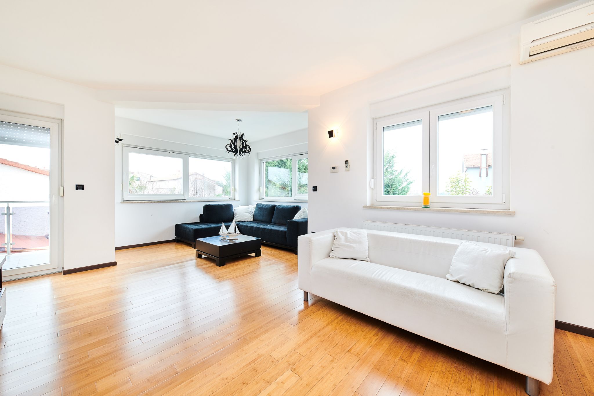 Uma sala de estar espaçosa com um sofá branco, um sofá escuro e uma mesa de centro. Há grandes janelas que proporcionam luz natural e vista para o exterior.