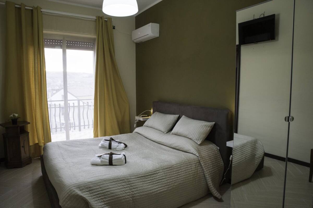 A hotel room with a large bed, a nightstand, a TV mounted on the wall, and a window with curtains.