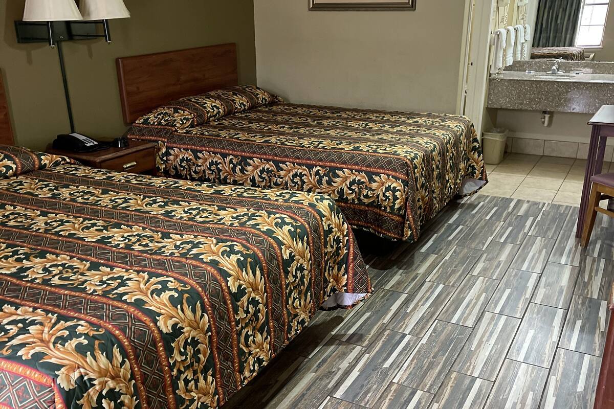 Quarto de hotel com três camas, cabeceira de madeira, mesa de cabeceira com abajur, quadro emoldurado na parede, banheiro com pia e espelho, e piso revestido de azulejos.