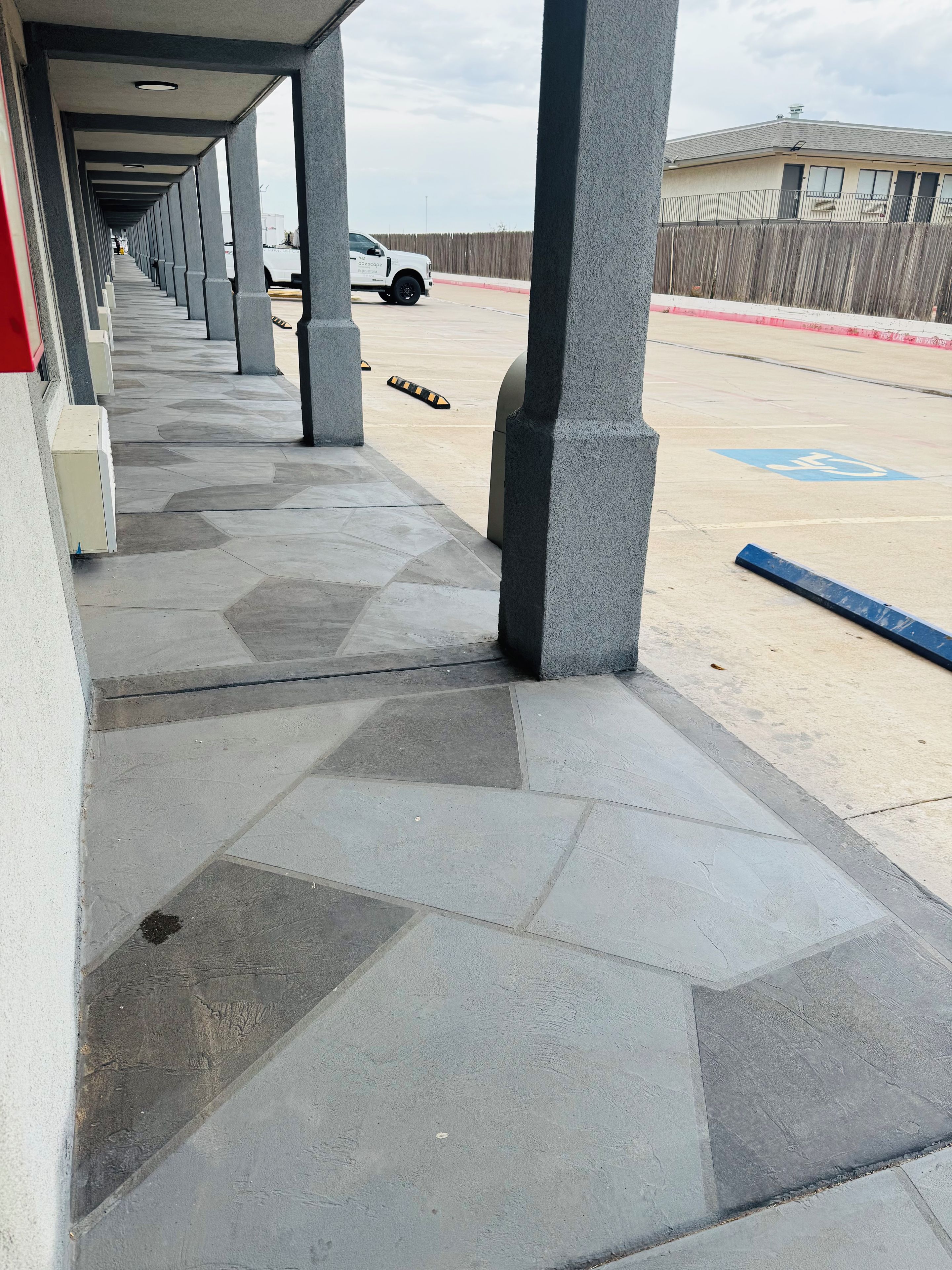 Un espacio al aire libre techado con piso de baldosas, columnas y un estacionamiento al fondo.