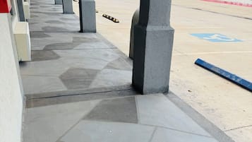Un espacio al aire libre techado con piso de baldosas, columnas y un estacionamiento al fondo.