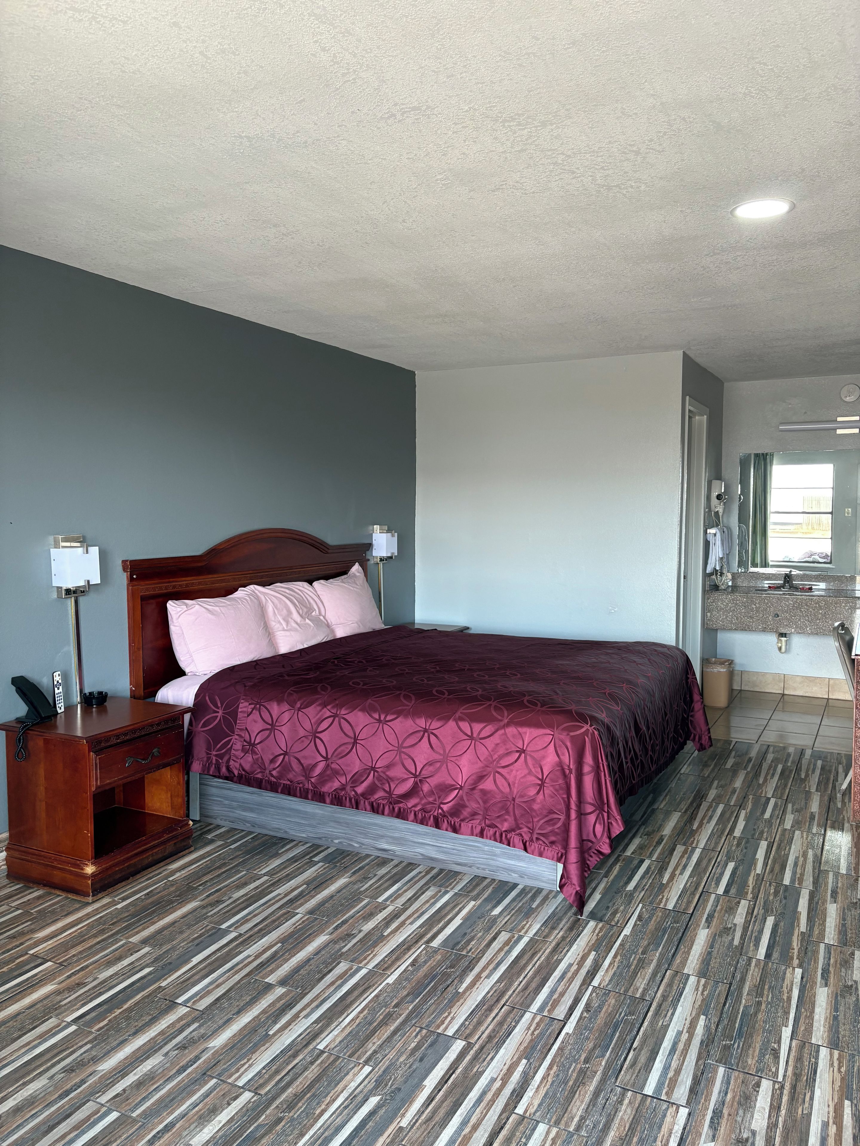 A hotel room with a bed, bedside table, lamp, and a bathroom visible through an open door.