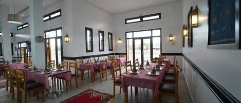 Un comedor con mesas dispuestas para comer, ventanas amplias y luces fijadas a la pared.