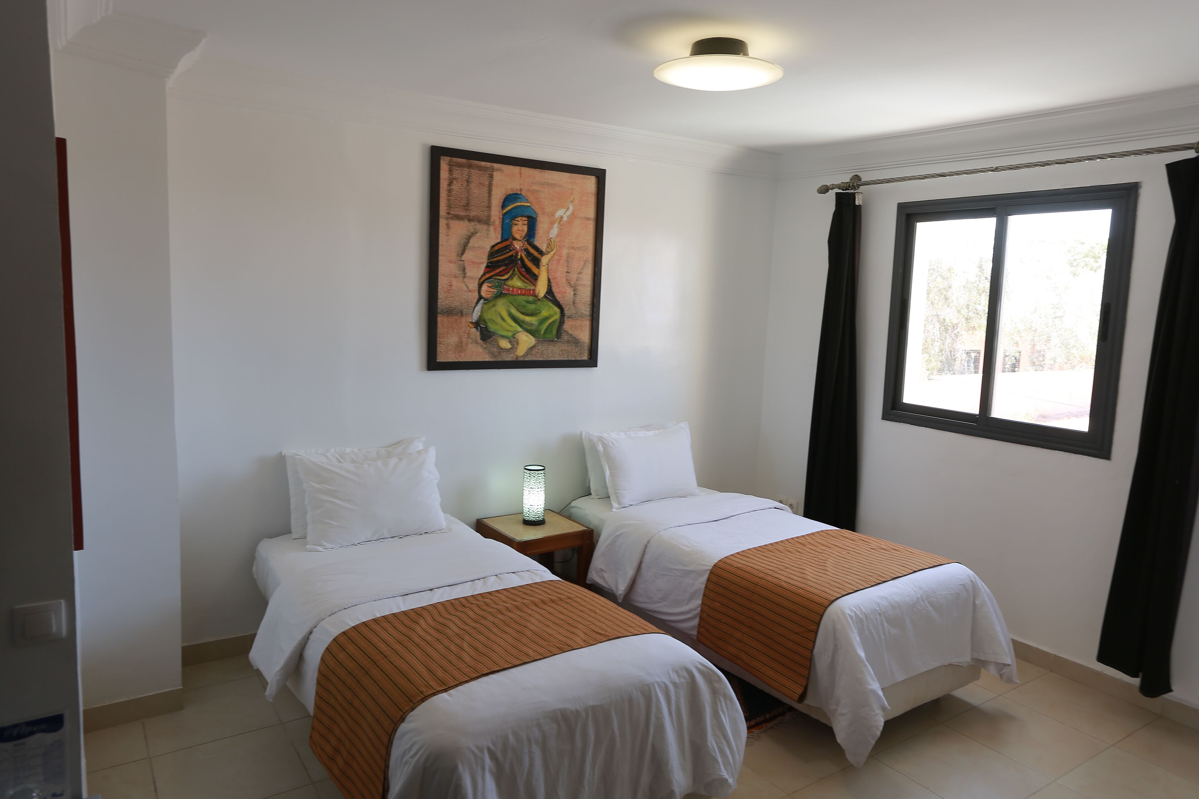 Habitación de hotel con dos camas individuales, una mesita de noche con lámpara, un cuadro en la pared y una ventana con cortinas.