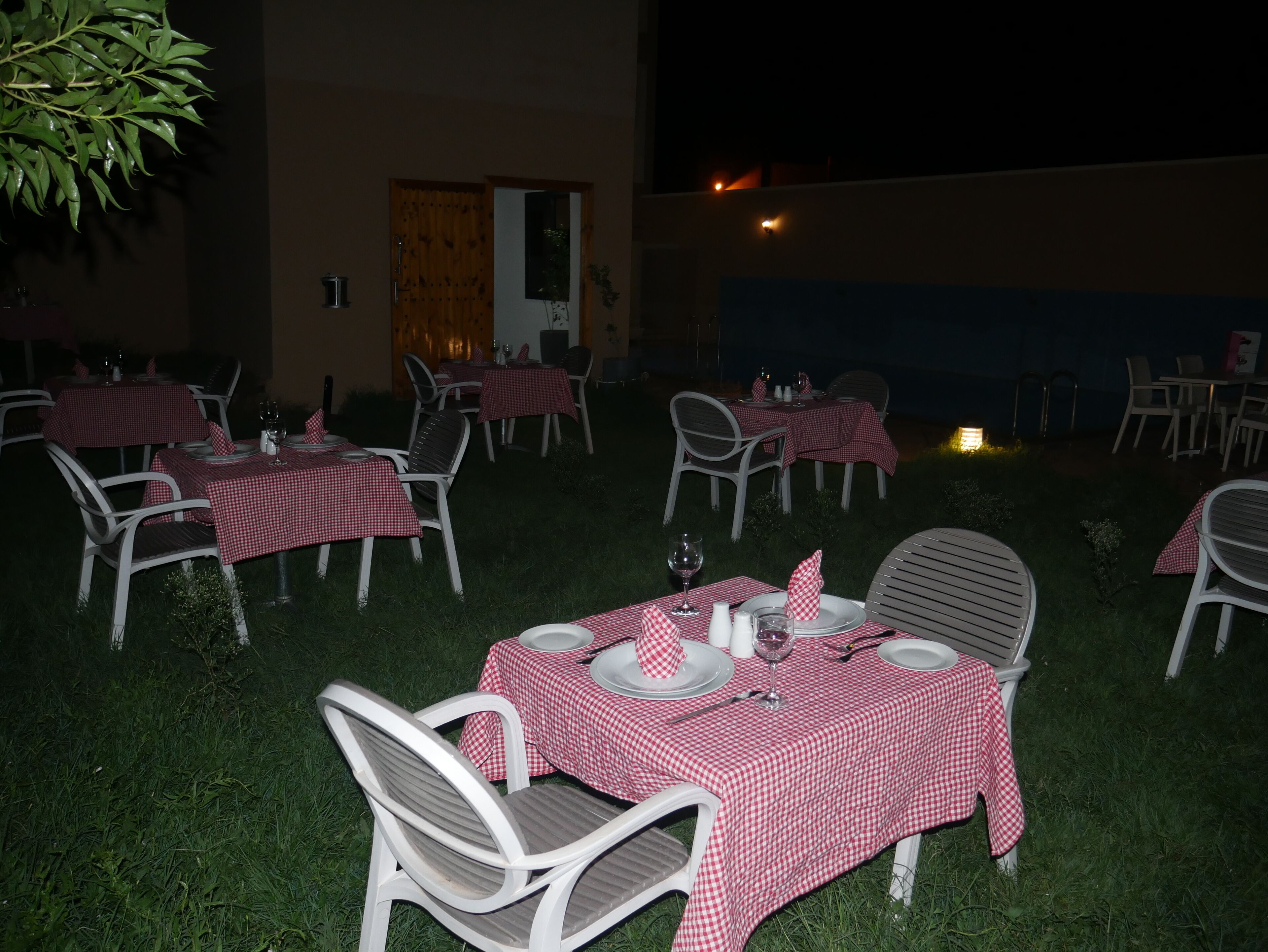 Un área de comedor al aire libre con mesas dispuestas para dos, cubiertas con manteles a cuadros rojos y blancos, rodeada de sillas blancas.