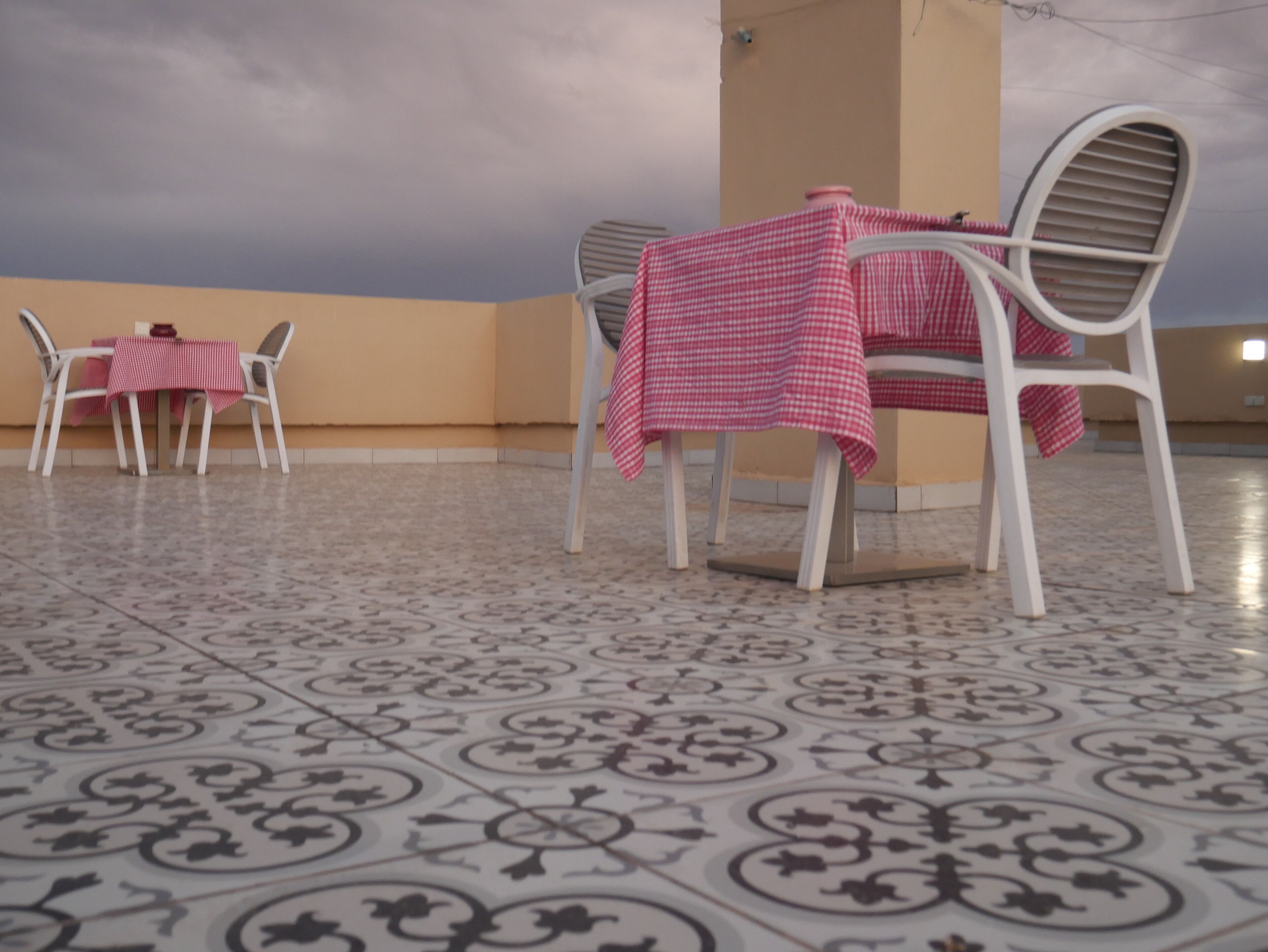 A terrace with a table and chairs covered with a checkered tablecloth, a tiled floor, and a cloudy sky in the background.