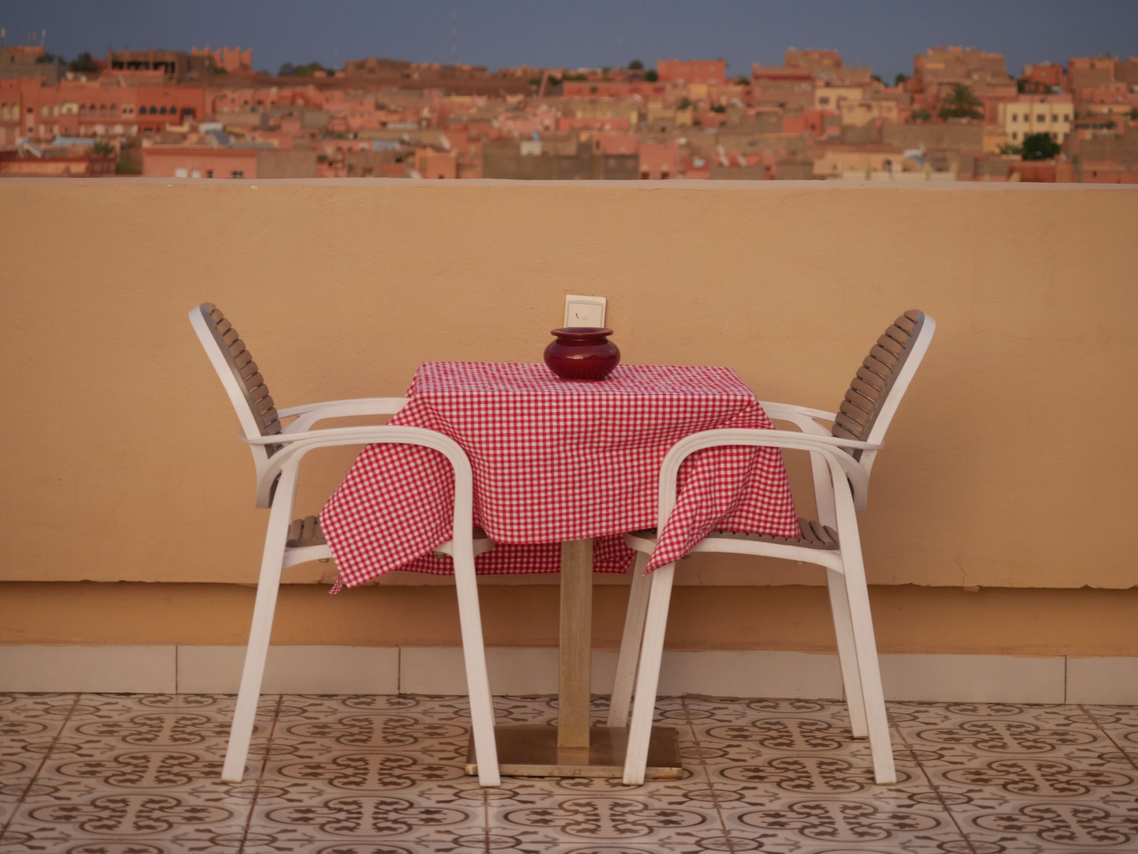 A table with a red and white checkered tablecloth, two white chairs, and a decorative bowl on the table. The background features a cityscape with buildings.