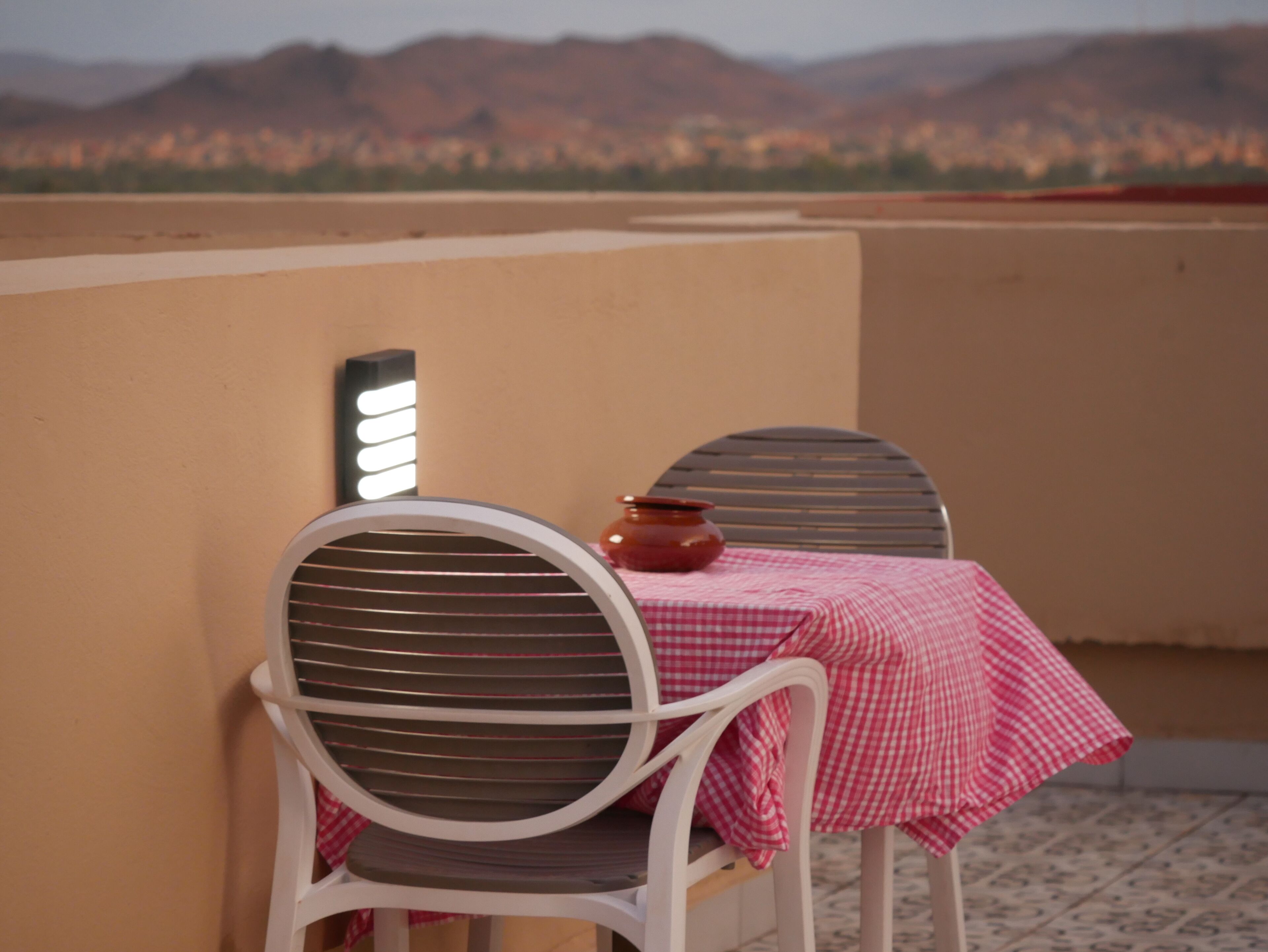 Un balcon avec deux chaises et une table, un lampadaire fixé au mur, et une vue sur des montagnes lointaines.