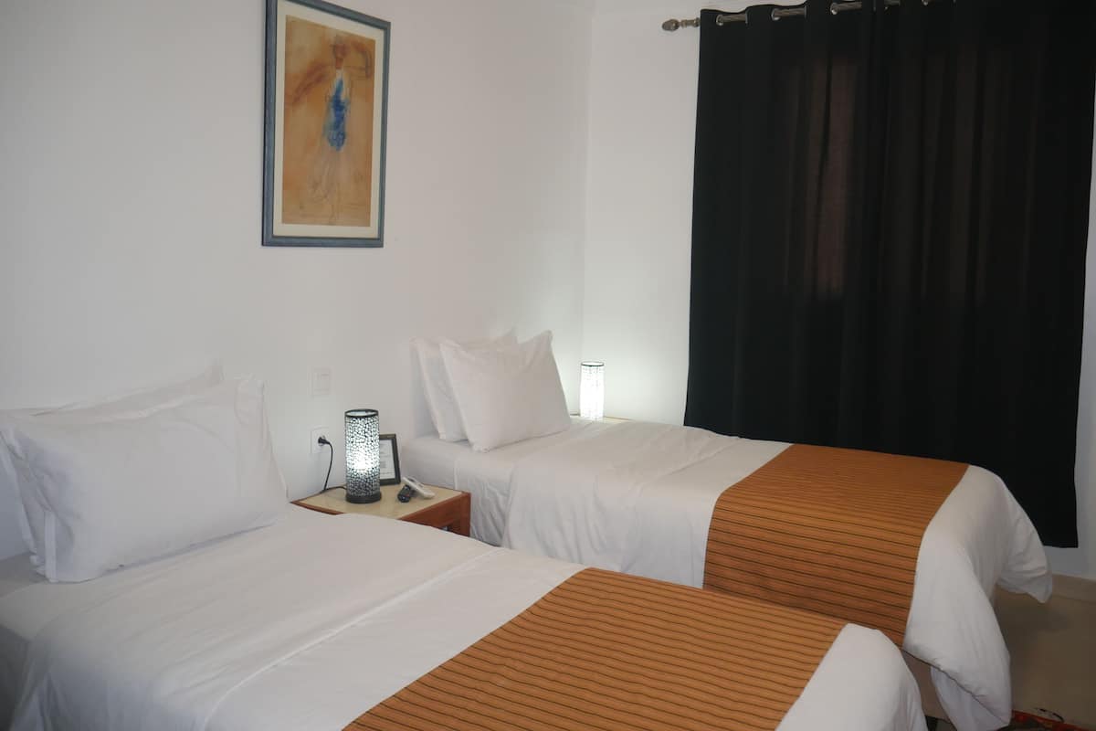 A hotel room with two beds, a nightstand with a lamp, a framed picture on the wall, and a black curtain.