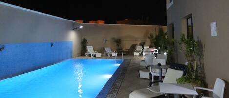 An outdoor swimming pool with lounge chairs and tables, surrounded by a building with a tiled floor.