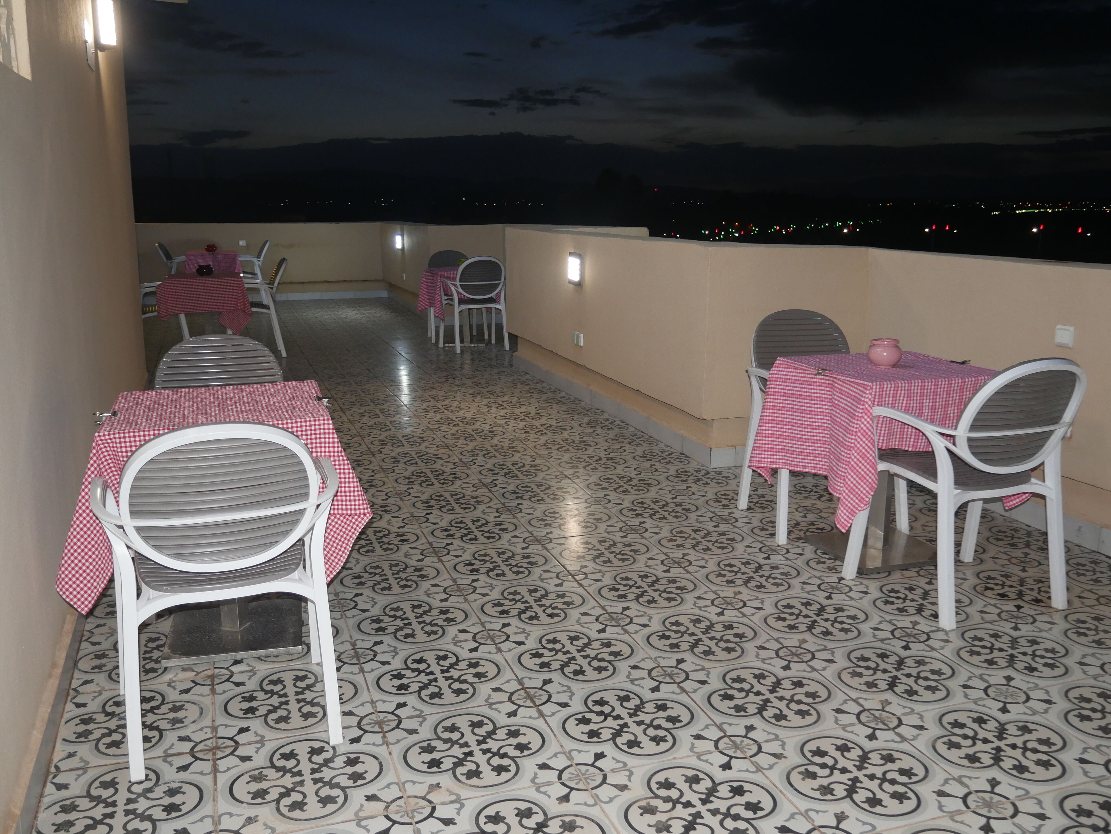 A terrace with tables and chairs, patterned floor tiles, and a view of the city at night.