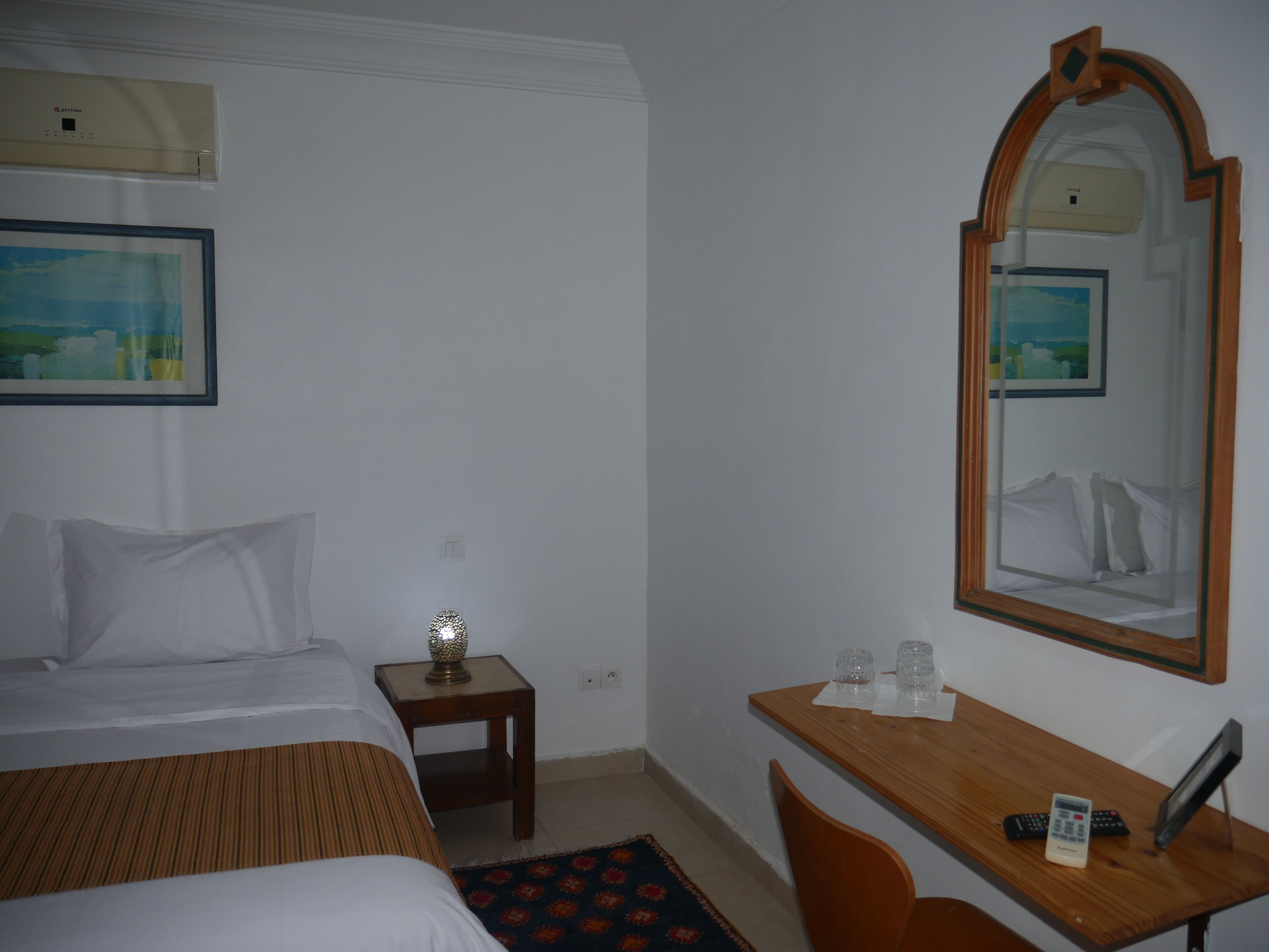 Habitación de hotel con cama, mesita de noche con lámpara, escritorio con silla, espejo y ventana.