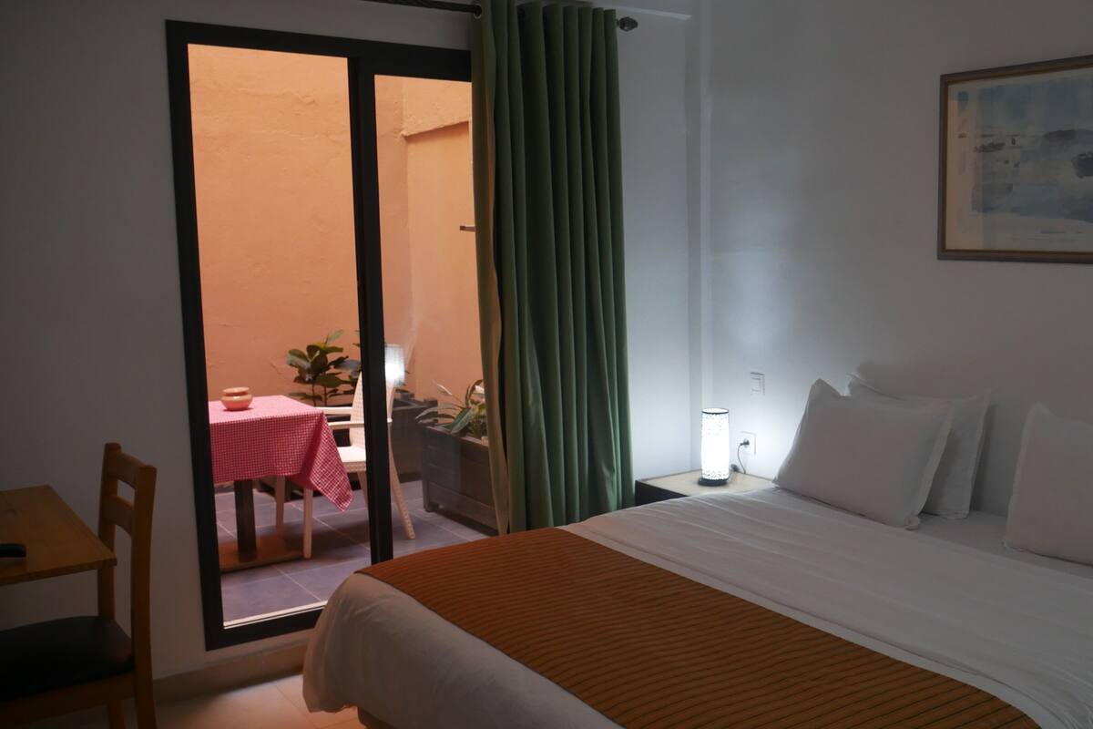 A hotel room with a bed, a table with a red tablecloth, a chair, and a view of an outdoor area with a table and chairs.