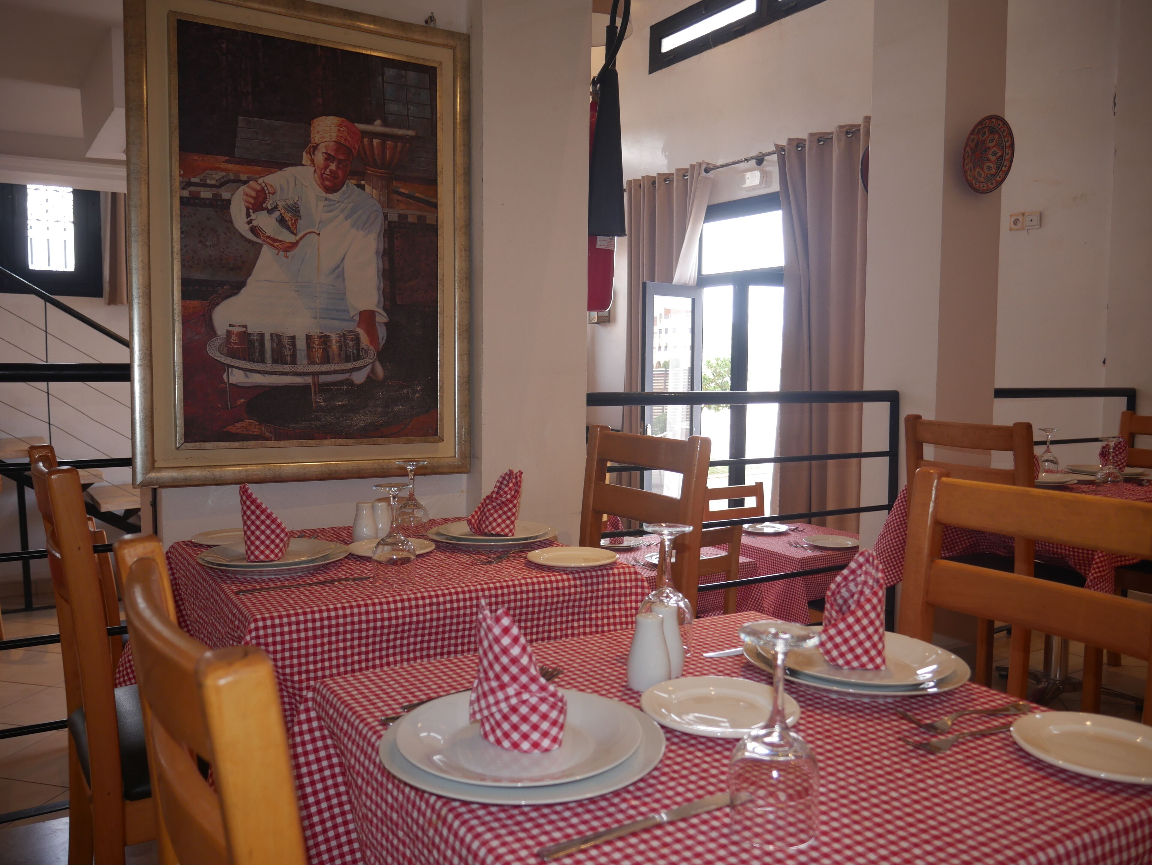 A dining area with red and white checkered tablecloths, wooden chairs, and a painting on the wall.