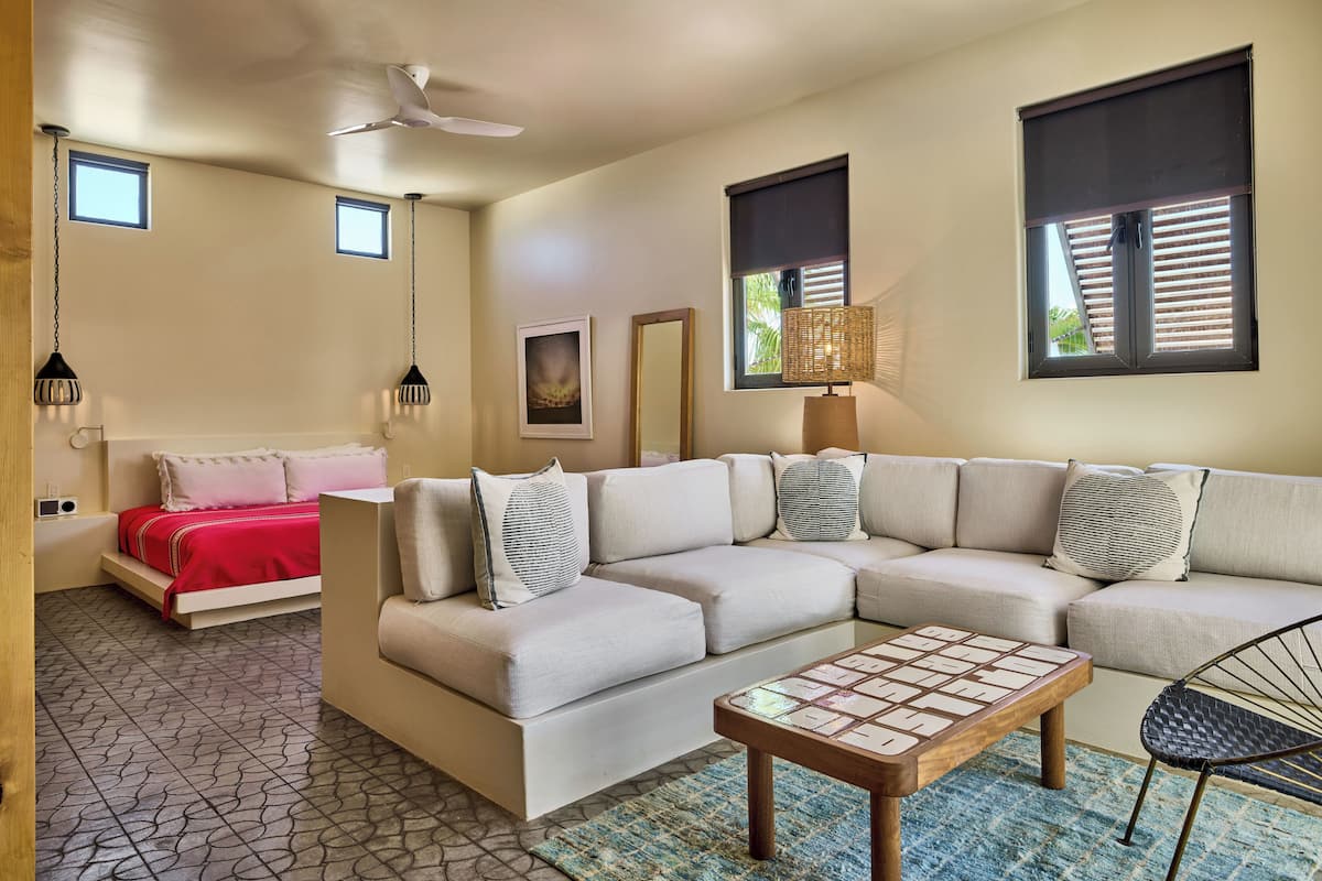 A modern living room with a sectional sofa, a coffee table, a bed with a red blanket, and a ceiling fan.