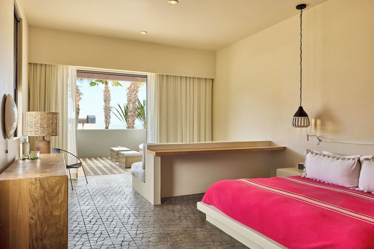 A hotel room with a bed, a desk, a chair, a lamp, and a view of palm trees through the window.