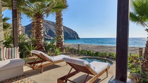 Un salón junto a la playa con dos tumbonas de madera, una piscina pequeña y vistas al océano y a una montaña.