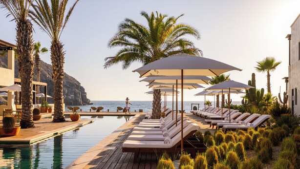 Zona junto a la piscina con sillones de descanso y sombrillas, palmeras y vista al mar.