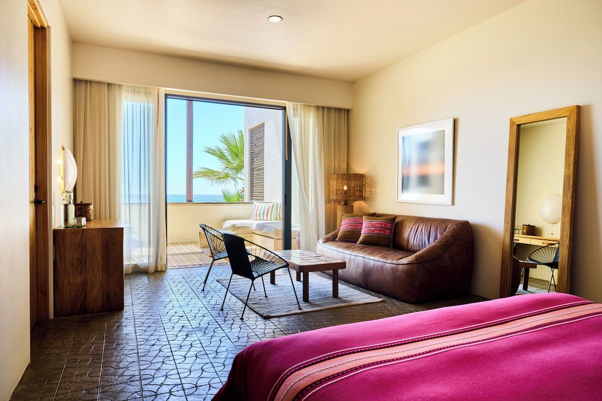 A hotel room with a large bed, a sofa, a desk, and a chair. There is a window with curtains and a view of palm trees.
