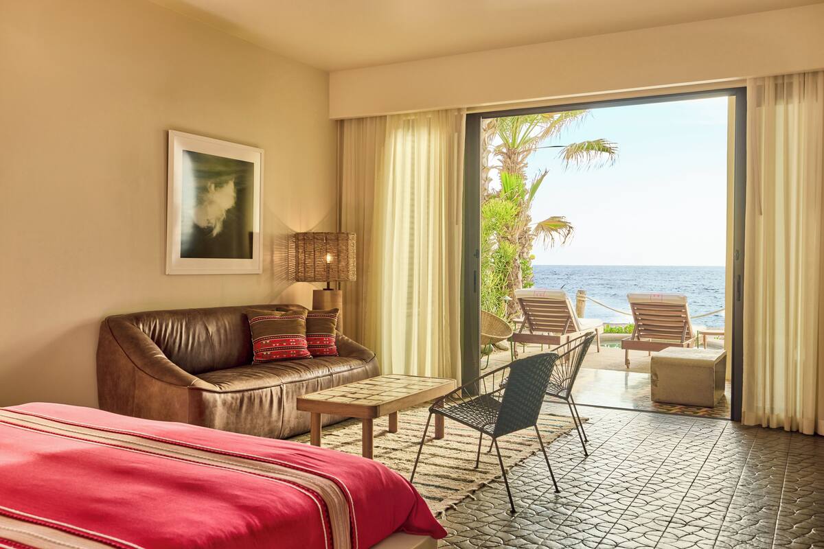 A hotel room with a leather sofa, a bed, a small table, and a chair. There is a large window with a view of the ocean and palm trees.