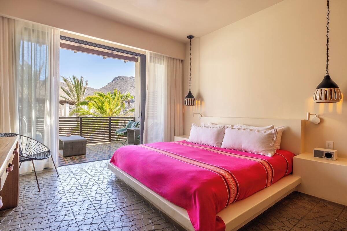 A hotel room with a large bed, a desk, and a chair. There is a balcony with a view of palm trees and mountains.