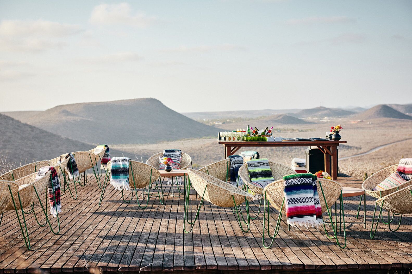 A rustic outdoor seating area with wicker chairs and a wooden table set up on a wooden deck, overlooking a mountainous landscape.