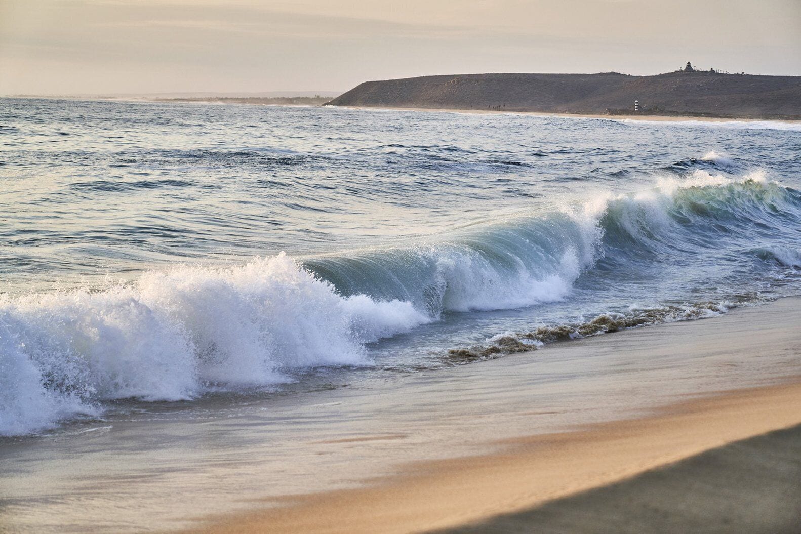 Ein Strand mit brechenden Wellen, feiner Sand und eine Hügelkette in der Ferne mit einem Bauwerk auf der Spitze.