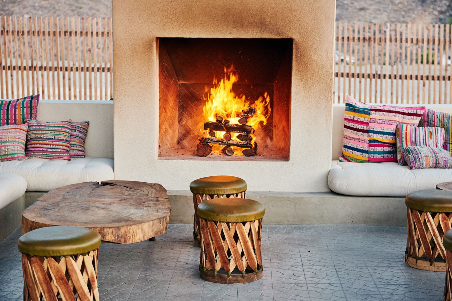 A cozy outdoor seating area with a built-in fire feature, cushioned sofas, and wooden stools.