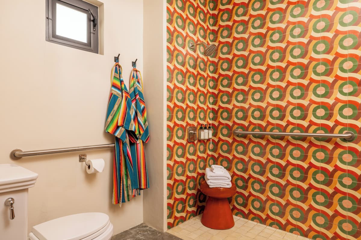 A bathroom with a toilet, a towel rack with colorful towels, a shower area with a patterned wall, and a window above the toilet.