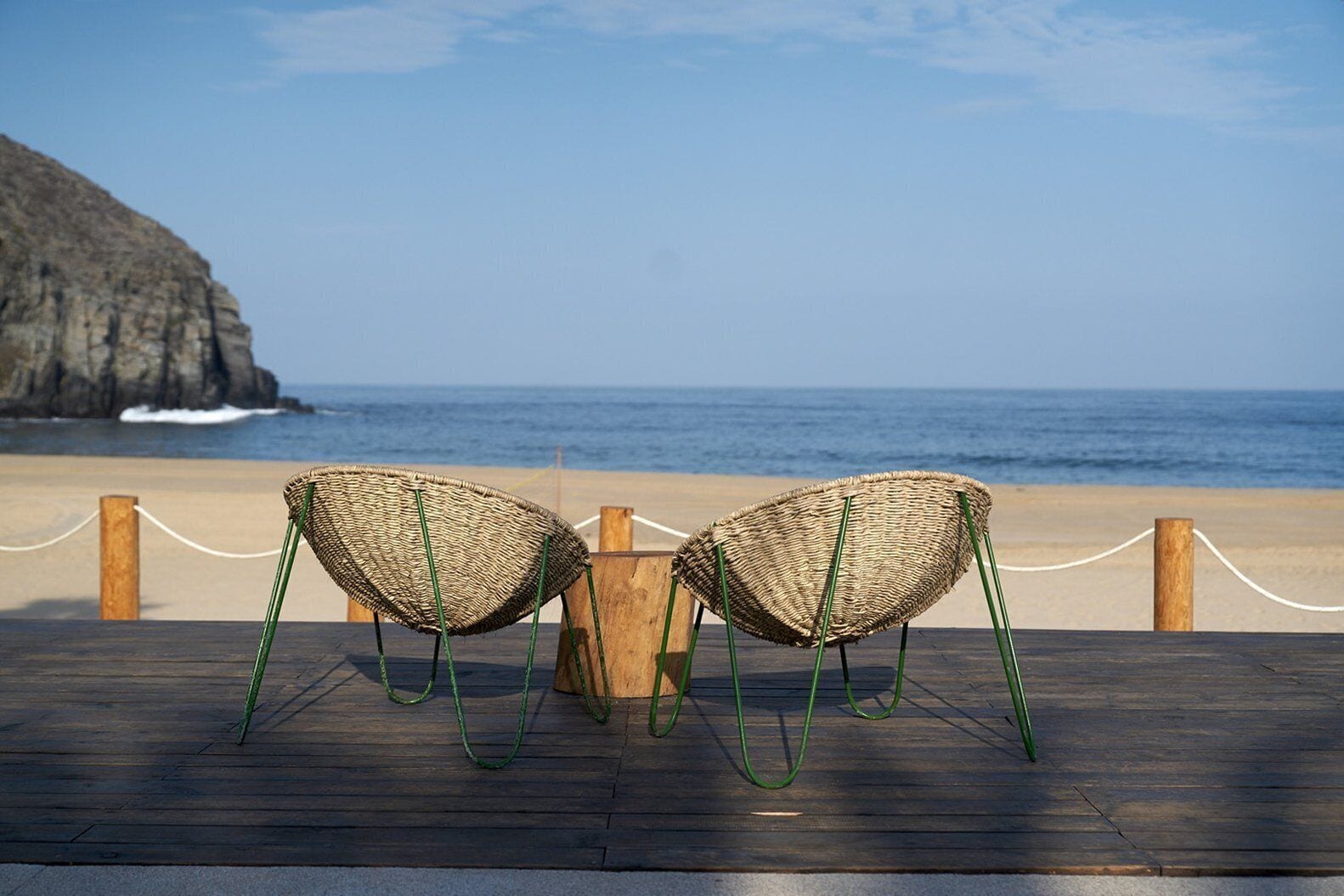 Two wicker chairs with green legs on a wooden deck overlooking a beach with a rocky cliff and the ocean.