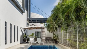 Un área de piscina moderna con piscina azul cristalina, sillones blancos para tomar el sol y una zona de estar cubierta.